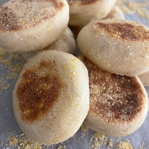 May include: A close-up of six golden brown English muffins on a white surface. The muffins are slightly toasted and have a crispy exterior.
