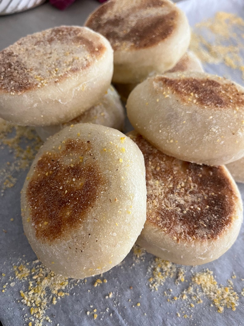 May include: A close-up of six golden brown English muffins on a white surface. The muffins are slightly toasted and have a crispy exterior.