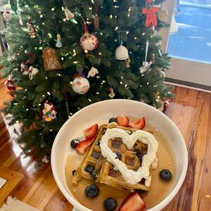 May include: Two waffles topped with whipped cream, blueberries, and strawberries. The whipped cream is shaped like a heart. The waffles are on a white plate with a brown wooden background.