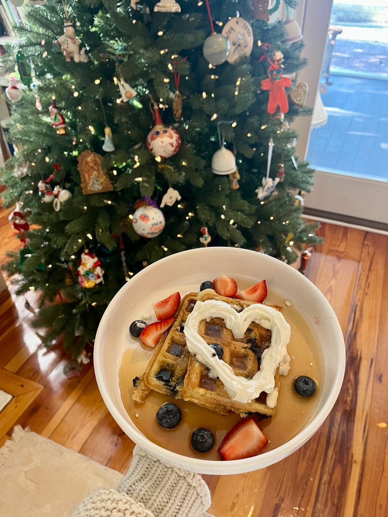 May include: Two waffles topped with whipped cream, blueberries, and strawberries. The whipped cream is shaped like a heart. The waffles are on a white plate with a brown wooden background.