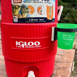 May include: Red Igloo 5-gallon Seat Top beverage cooler with a white lid and a white spigot. The cooler has a black cup holder attached to the side. A green cup with a logo is in the cup holder.