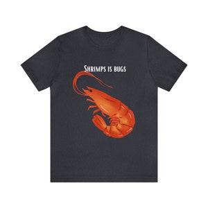 May include: Dark grey t-shirt with an orange shrimp illustration and the text "SHRIMPS IS BUGS" in white. The shrimp is the focal point of the design. The shirt is made of a soft material.
