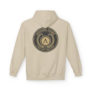 Może przedstawiać: Beżowa bluza z kapturem z okrągłym emblematem na plecach. Emblemat zawiera napisy "MOLON LABE" i "COME AND TAKE THEM" wokół centralnego wzoru z włócznią i grecką literą lambda.