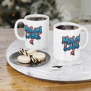 May include: Two white ceramic mugs with the text "MOLON LABE" in blue and red graffiti-style lettering. Each mug contains dark liquid and is placed on a marble tray. A plate of macarons and a spoon are also on the tray.