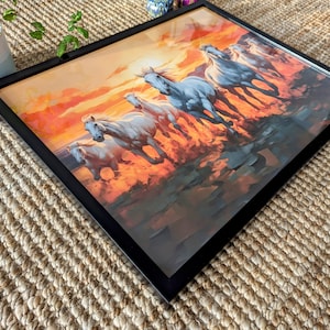 May include: A framed painting of a group of white horses running through a field at sunset. The horses are silhouetted against the orange and red sky. The painting is in a black frame.