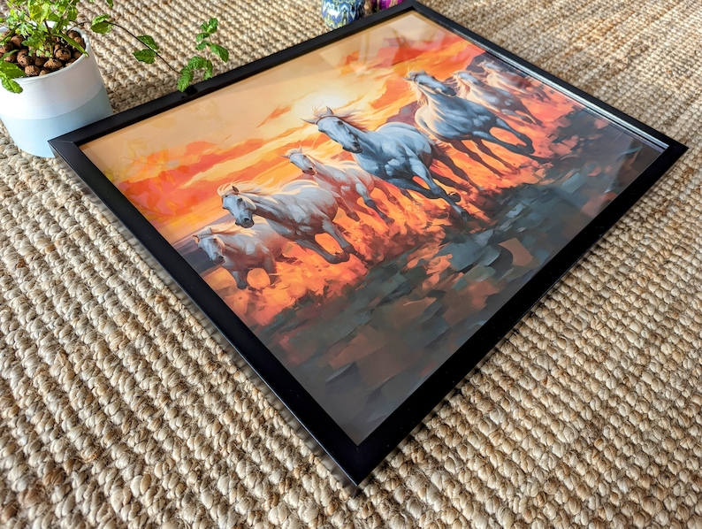 May include: A framed painting of a group of white horses running through a field at sunset. The horses are silhouetted against the orange and red sky. The painting is in a black frame.