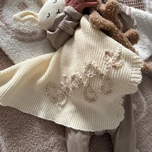 May include: A cream-colored knitted security blanket featuring a plush rabbit head with embroidered facial features and the name "Bodie" stitched in a textured yarn on the front. The blanket has a soft, garter stitch texture and a scalloped edge.