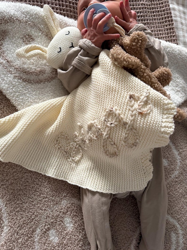 May include: A cream-colored knitted security blanket featuring a plush rabbit head with embroidered facial features and the name "Bodie" stitched in a textured yarn on the front. The blanket has a soft, garter stitch texture and a scalloped edge.