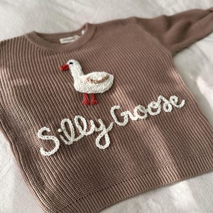 Könnte beinhalten: Ein brauner Strickpullover mit einem weißen Gänse-Applikation und den Worten "Silly Goose" in weißer Schrift. Die Gans hat einen weißen Körper, einen beigen Flügel und rote Füße und einen Schnabel. Der Pullover hat lange Ärmel.