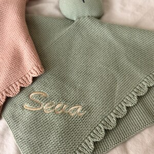 May include: A sage green knitted security blanket featuring a rabbit head with embroidered closed eyes and a nose. The blanket has a ruffled edge and the name "Sena" embroidered in light beige thread. A partial pink knitted blanket is visible.