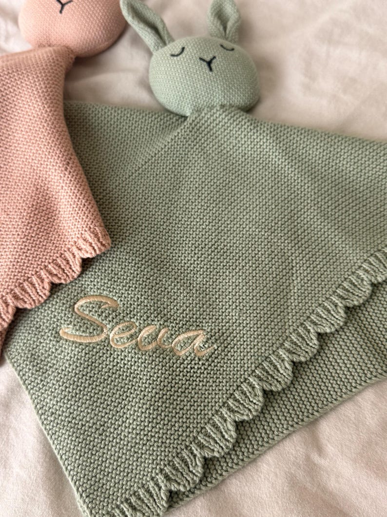 May include: A sage green knitted security blanket featuring a rabbit head with embroidered closed eyes and a nose. The blanket has a ruffled edge and the name "Sena" embroidered in light beige thread. A partial pink knitted blanket is visible.