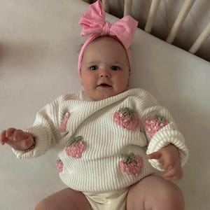 Peut inclure: Un bébé portant un pull en tricot blanc avec des fraises brodées roses et un bandeau rose en forme de nœud. Le bébé est allongé sur un drap blanc dans un berceau.
