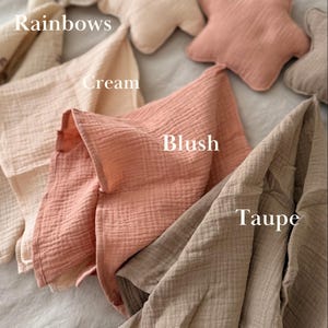 May include: A collection of soft muslin cloths and star-shaped pillows in various colors labeled as Rainbows, Cream, Blush, and Taupe. The fabric has a crinkled texture, and the items are arranged in a layered display showing the different shades.
