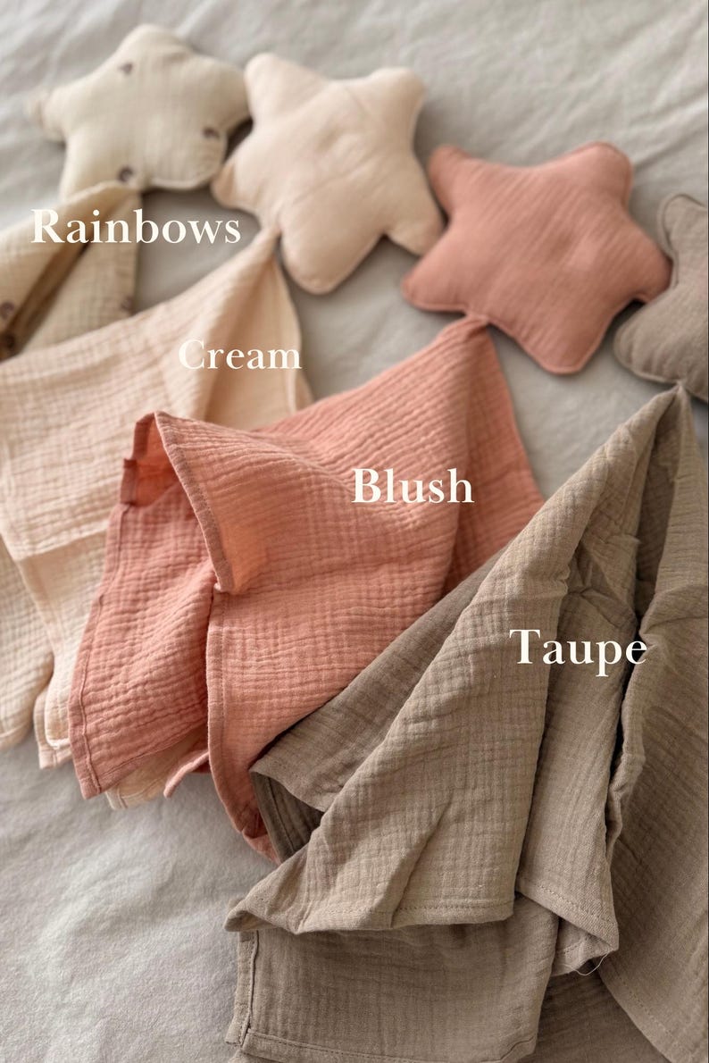 May include: A collection of soft muslin cloths and star-shaped pillows in various colors labeled as Rainbows, Cream, Blush, and Taupe. The fabric has a crinkled texture, and the items are arranged in a layered display showing the different shades.