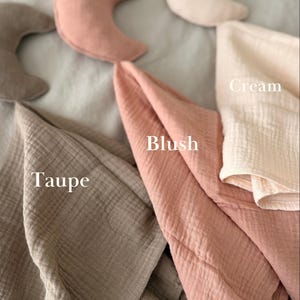 May include: A collection of crescent moon-shaped pillows and folded muslin fabric pieces in cream, blush, and taupe colors. The text labels Cream, Blush, and Taupe are printed on the image to identify the specific color of each fabric item.