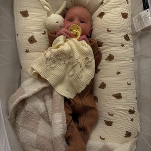 May include: A baby wearing a brown ribbed footed sleeper with a yellow pacifier, lying in a cream-colored baby nest with a brown bear and moon print. The baby holds a cream-colored knitted security blanket and a small white crocheted rabbit toy.
