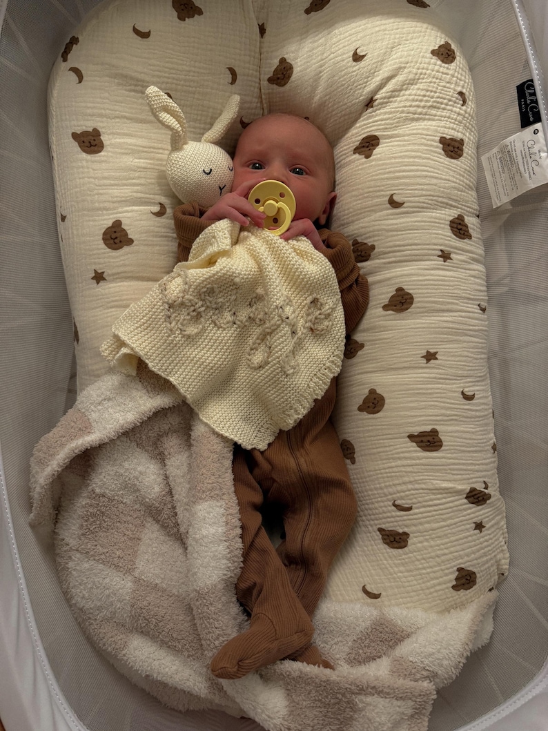 May include: A baby wearing a brown ribbed footed sleeper with a yellow pacifier, lying in a cream-colored baby nest with a brown bear and moon print. The baby holds a cream-colored knitted security blanket and a small white crocheted rabbit toy.