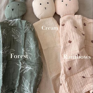 May include: Three soft muslin security blankets with rabbit heads. The left one is forest green with white leaf patterns. The middle one is solid cream. The right one is light pink with small brown rainbow prints. Each has knotted corners.