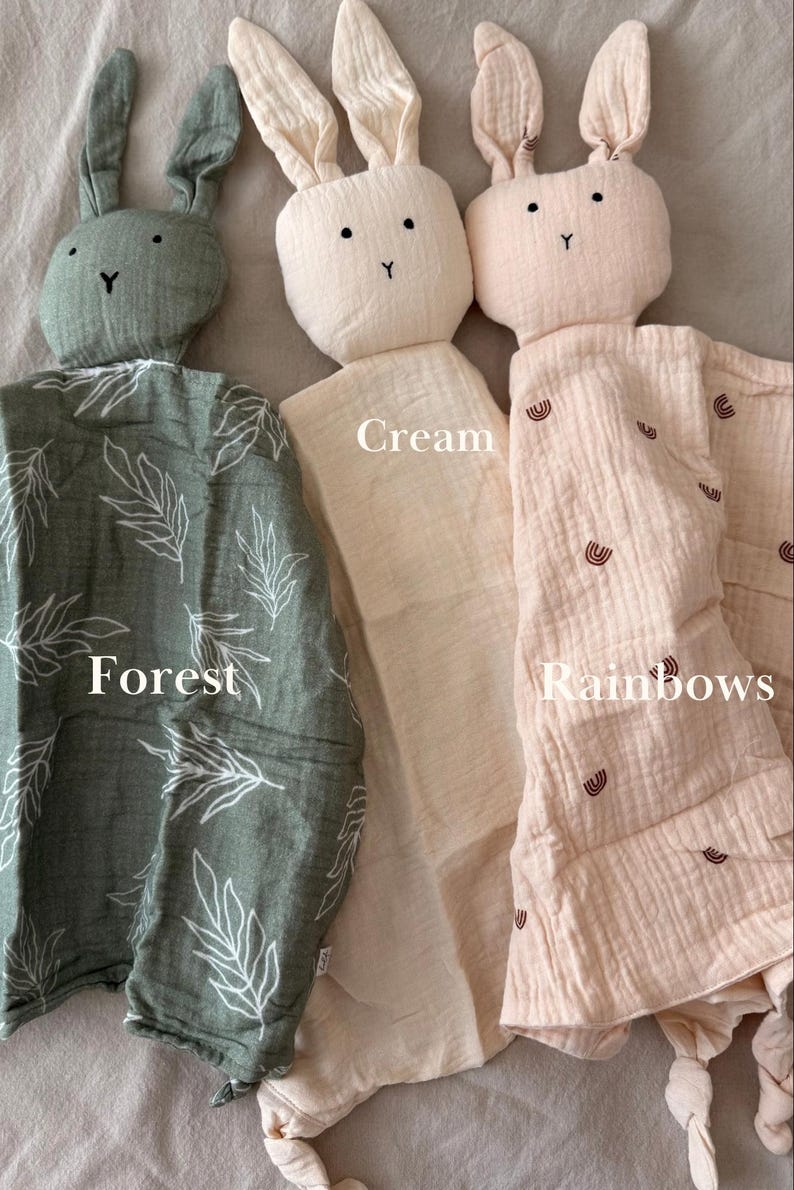 May include: Three soft muslin security blankets with rabbit heads. The left one is forest green with white leaf patterns. The middle one is solid cream. The right one is light pink with small brown rainbow prints. Each has knotted corners.