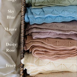 May include: A stack of six knitted blankets in a woven basket, labeled with the colors Sage, Sky Blue, Mauve, Dusty Rose, Ivory, and Speckled Oat. Each blanket features a textured knit pattern with a scalloped edge design along the border.