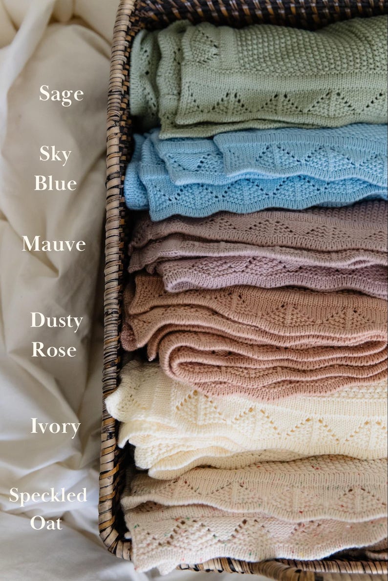 May include: A stack of six knitted blankets in a woven basket, labeled with the colors Sage, Sky Blue, Mauve, Dusty Rose, Ivory, and Speckled Oat. Each blanket features a textured knit pattern with a scalloped edge design along the border.