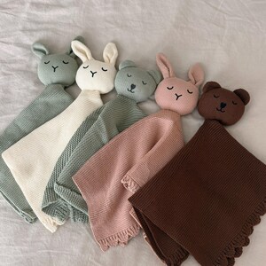 May include: Five knitted security blankets featuring plush animal heads. The collection includes two rabbits and three bears in sage green, cream, olive green, dusty pink, and dark brown. Each blanket has a textured knit body and a scalloped edge.