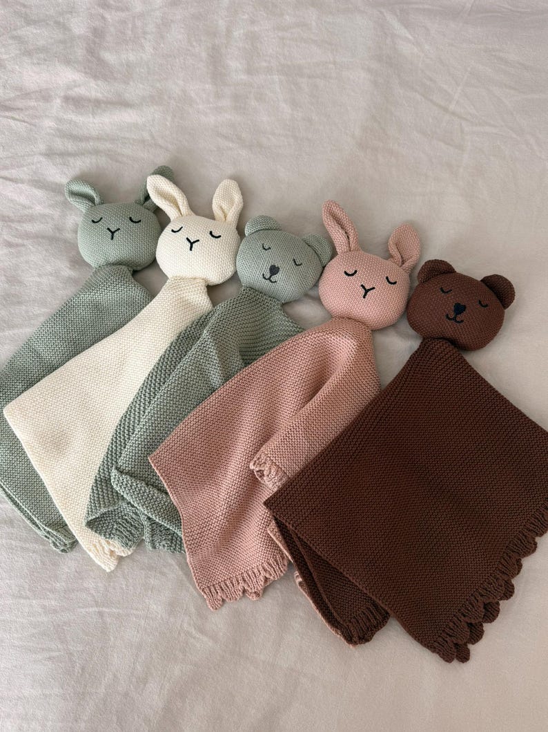 May include: Five knitted security blankets featuring plush animal heads. The collection includes two rabbits and three bears in sage green, cream, olive green, dusty pink, and dark brown. Each blanket has a textured knit body and a scalloped edge.