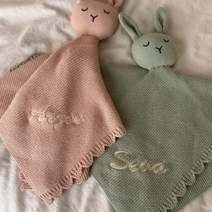 May include: Two knitted security blankets with rabbit heads, one in dusty pink and one in sage green. Each blanket features a textured knit pattern, a fringed edge, and embroidered names, Arzae on the pink blanket and Seva on the green one.
