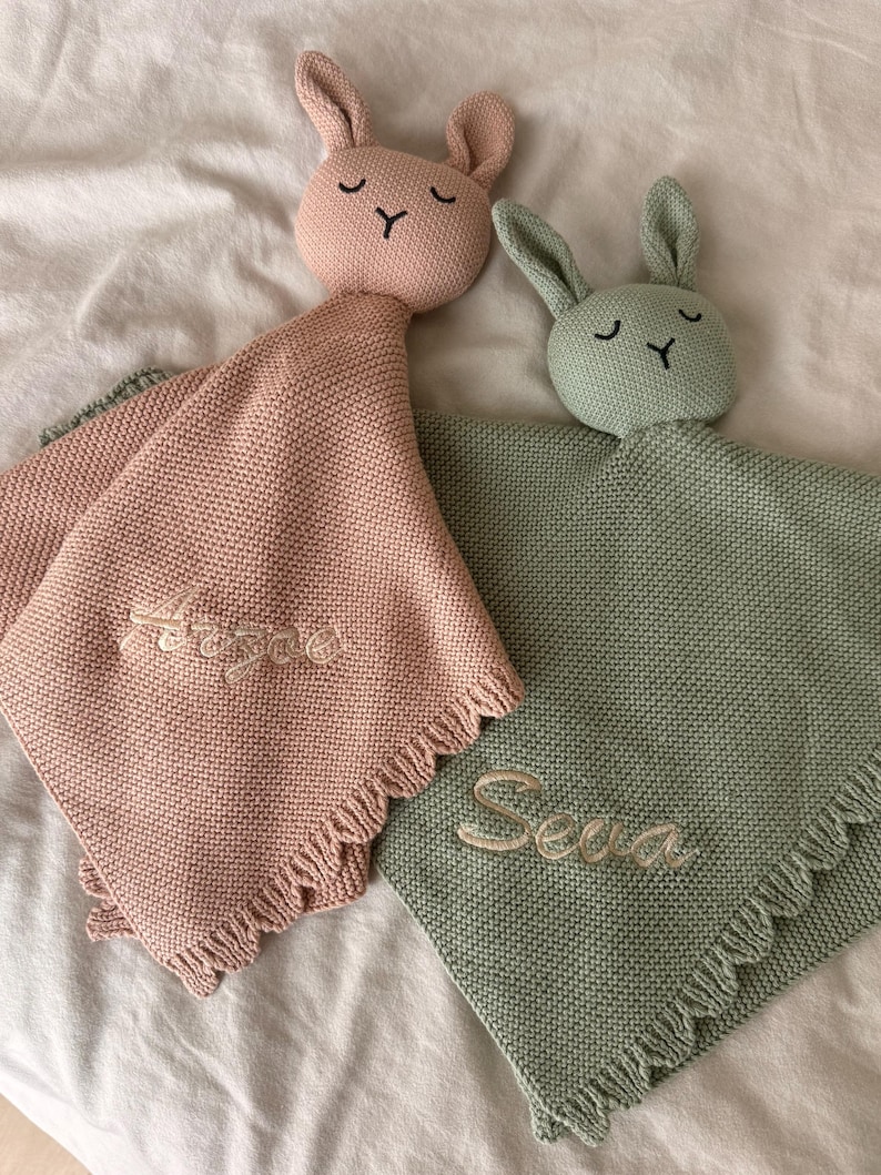 May include: Two knitted security blankets with rabbit heads, one in dusty pink and one in sage green. Each blanket features a textured knit pattern, a fringed edge, and embroidered names, Arzae on the pink blanket and Seva on the green one.