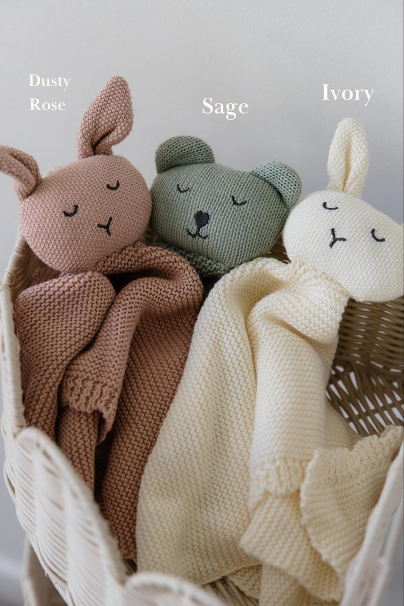 May include: Three knitted security blankets featuring animal heads in dusty rose, sage, and ivory colors. Each blanket has embroidered closed eyes and a nose. The dusty rose and ivory blankets have rabbit ears, while the sage blanket has bear ears.