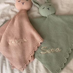 May include: Two knitted security blankets shaped like bunnies, one in dusty pink and one in sage green. Each features a bunny head with embroidered eyes and nose, a textured knit body, and a fringed edge. The pink blanket is embroidered with the name Arzae, and the green blanket is embroidered with the name Seva.
