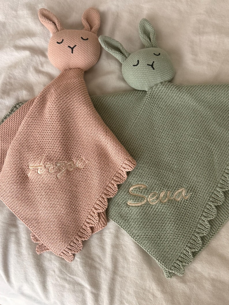 May include: Two knitted security blankets shaped like bunnies, one in dusty pink and one in sage green. Each features a bunny head with embroidered eyes and nose, a textured knit body, and a fringed edge. The pink blanket is embroidered with the name Arzae, and the green blanket is embroidered with the name Seva.