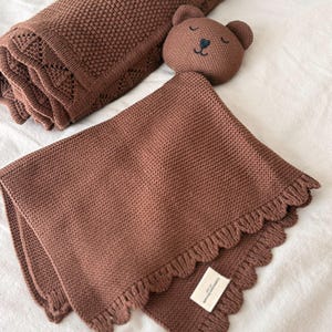 May include: A brown knitted security blanket featuring a plush bear head with embroidered facial features and a ruffled edge. The blanket has a textured knit pattern and includes a small white rectangular fabric label attached to the corner.