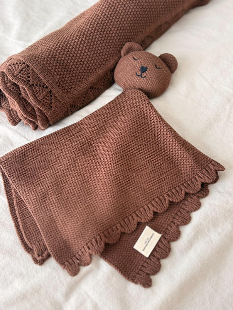 May include: A brown knitted security blanket featuring a plush bear head with embroidered facial features and a ruffled edge. The blanket has a textured knit pattern and includes a small white rectangular fabric label attached to the corner.