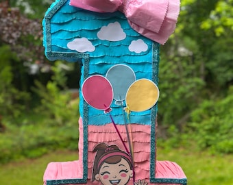 May include: A turquoise and pink number one piñata with a pink bow, white clouds, and a girl with brown hair and a blue dress holding balloons. The piñata is decorated with glitter.