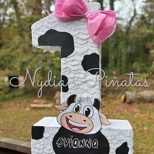 May include: A white and black cow-themed piñata shaped like the number one. The piñata has a pink bow on top and a cartoon cow face on the front. The name "Evianna" is written on the front of the piñata.