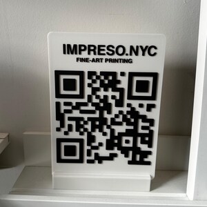 May include: A white rectangular sign with a black QR code and the text "IMPRESO.NYC FINE-ART PRINTING." The sign is displayed on a white shelf. The QR code is a square pattern of black and white squares.