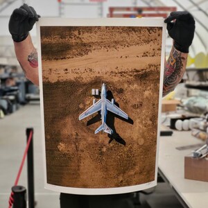 May include: An aerial photograph of a silver airplane on a dirt field. The airplane is facing the camera and has its wings spread out. The photo is printed on a white paper and is being held by a person.