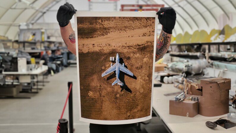 May include: An aerial photograph of a silver airplane on a dirt field. The airplane is facing the camera and has its wings spread out. The photo is printed on a white paper and is being held by a person.