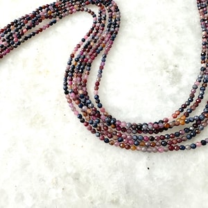 May include: A strand of multi-colored gemstone beads. The beads are small and round, and they are arranged in a long, flowing strand. The colors of the beads are shades of blue, purple, and pink.