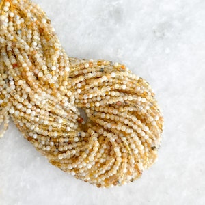 May include: A strand of small, round, yellow and white opal beads. The beads are strung on a thin, clear thread. The beads are all approximately the same size and shape.