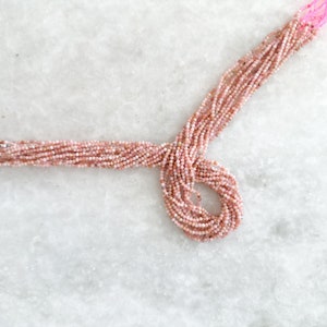 May include: A strand of pink opal beads, approximately 2mm in diameter, arranged in a loose circle on a white marble surface.