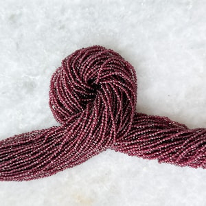 May include: A strand of deep red garnet beads, approximately 2mm in size, arranged in a loose coil on a white marble surface.