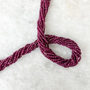 May include: A strand of deep red gemstone beads, likely ruby or garnet, arranged in a loose circle on a white marble surface. The beads are faceted and have a smooth, polished finish.