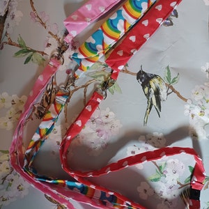 以下が含まれることがあります： ハートと虹のパターンが施されたカラフルなファブリック製のネックストラップが3本。各ネックストラップには金属製の留め具と黒いプラスチック製のクリップが付いています。