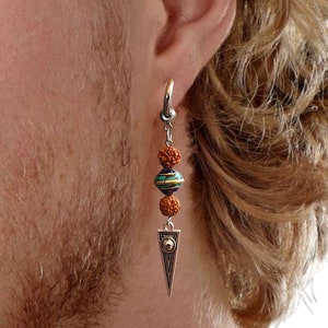 May include: A silver hoop earring with a dangling design. The earring features three beads in shades of brown and turquoise, and a triangular pendant with a geometric design. The earring is worn on the ear.