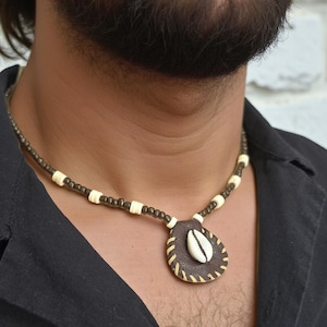 Puede incluir: Un collar de cuentas marrón y crema con un colgante. El colgante es una forma de lágrima de cuero marrón con una concha de color crema en el centro. El collar lo lleva una persona que lleva una camisa negra.