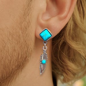 Può includere: Un orecchino d'argento con una pietra turchese quadrata, collegato a una piuma pendente con una pietra turchese più piccola. L'orecchino è indossato su un orecchio.