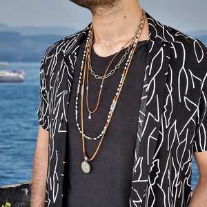 May include: A man wearing a black and white patterned shirt with a layered necklace featuring a round pendant. The necklace includes brown leather cord, silver chain, and beaded strands.