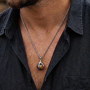 Può includere: Collana color argento con un ciondolo con una pietra rotonda color ambra. Il ciondolo è tenuto in un'incastonatura a forma di artiglio. La catena ha un design a corda. La persona indossa una camicia blu scuro.